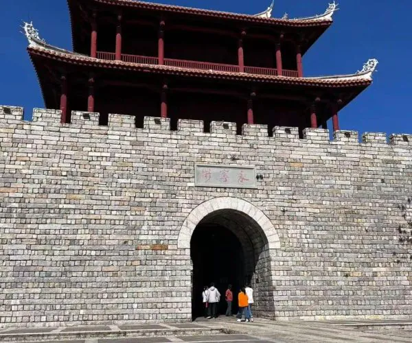 Shishi Ancient Town,Quanzhou,Chinatravel Stone fortress gate with an arched entrance and a red, multi-story pavilion atop, set against a blue sky.