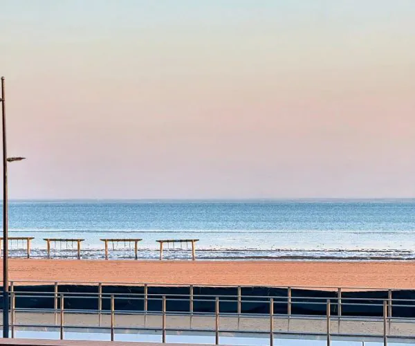 Qinhuangdao Weather,Qinhuangdao,Chinatravel Serene beach scene with a pastel sky, calm blue ocean, and empty benches along the shore behind a railing.
