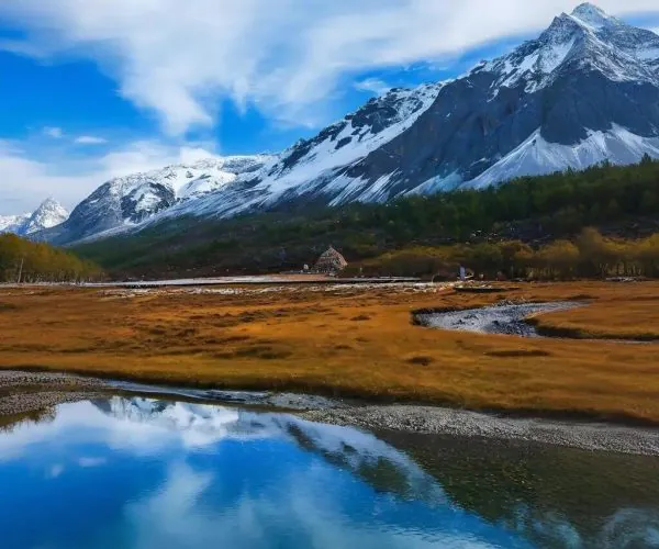 Daocheng & Yading ,Westren Sichuan,Chinatravel Snow-capped mountains beside a calm reflective lake under a blue sky with wispy clouds and a grassy foreground.