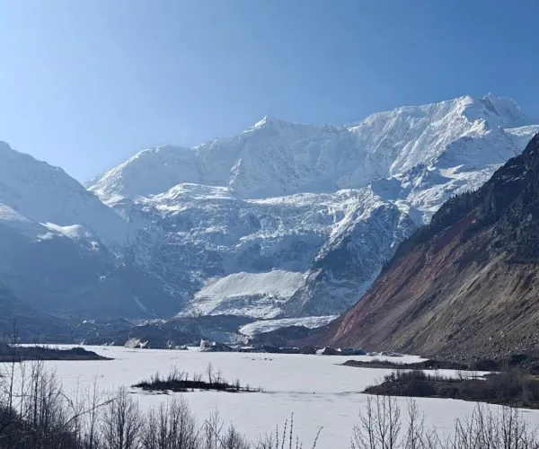 Midui Glacier,Nyingchi,China tour Snowy mountain range under a clear blue sky, with a frozen lake in the valley and bare shrubs in the foreground.