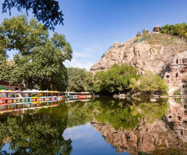 Red Hill Park,Urumqi,Chinatrip Row of colorful paddle boats docked along a tree-lined lakeshore with a rocky hill and pavilion above, reflecting in calm water.