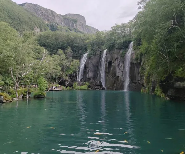 Green Deep Pool,Baishan,Chinatrip Turquoise lake with three waterfalls cascading down rocky cliffs, surrounded by lush trees and a wooden viewing deck on the left.
