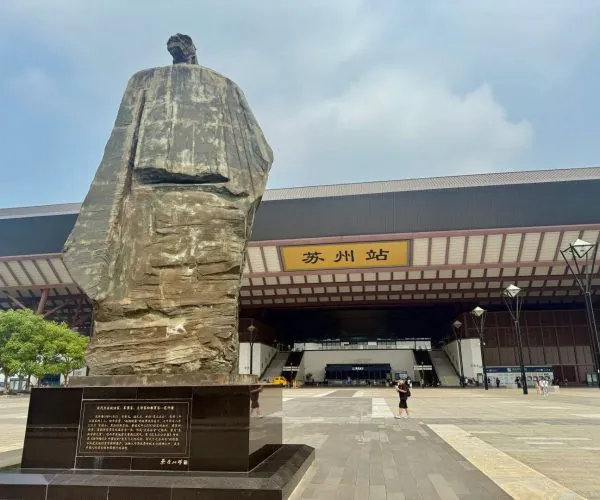 Suzhou Railway Station,Suzhou,Transportation Large rock statue on a dark pedestal in front of Suzhou Station, with a wide modern station entrance and trees nearby.