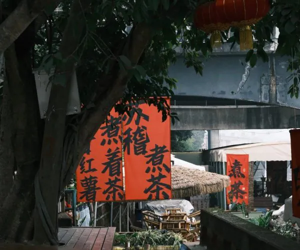 Suji Ancient Town,Leshan,Chinatravel Orange banners with black Chinese characters hang beside a tree in a shaded outdoor market, with wooden benches and market stalls in the background.