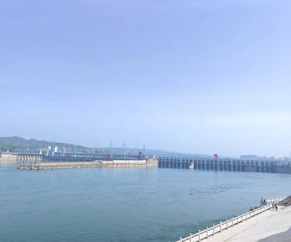 Gezhouba Water Conservancy,Yichang,Chinatravel Wide river with a long concrete dam or bridge spanning the water, and a waterfront walkway on the right where a few people stroll under a clear blue sky