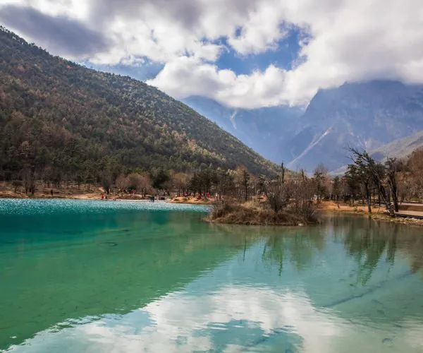 Blue Moon Valley,Lijang,Chinatravel Turquoise lake with reflected clouds, tree-covered hills on the left and misty mountains in the background; people gather on the sandy shore.