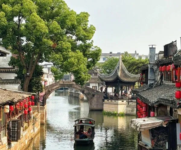Xitang Water Town,Hangzhou,Chinatravel Canal scene in a historic Chinese town with a small boat on calm water, stone bridge, pagoda-style pavilion, red lanterns along the banks, and green trees in the background.