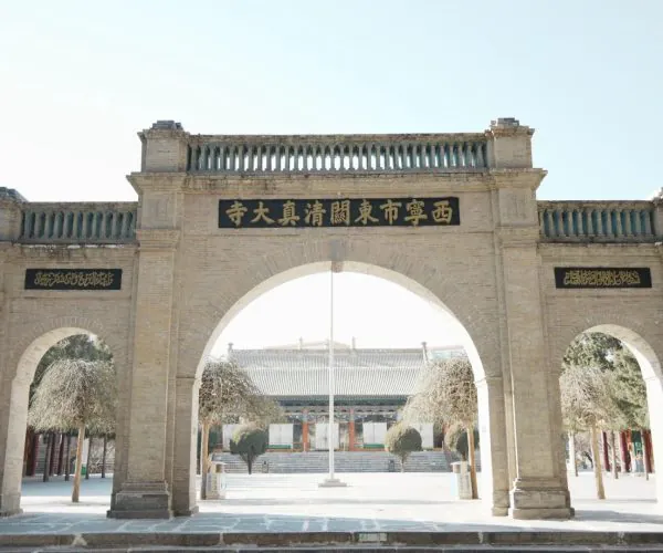 Dongguan Mosque,Xining,Chinatrip Stone city/temple entrance arch made of beige brick, with a central black plaque featuring gold Chinese characters above the arch; courtyard beyond with trees and a long building visible in distance.