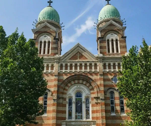Facade of a Byzantine-style church with two green domes and brick arches, flanked by trees.