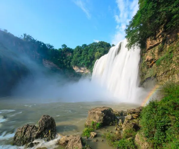 Guiyang Wearther,Guiyang,Chinatrip Waterfall pouring over a rocky cliff into a misty pool with a small rainbow near the right edge and lush green trees surrounding.