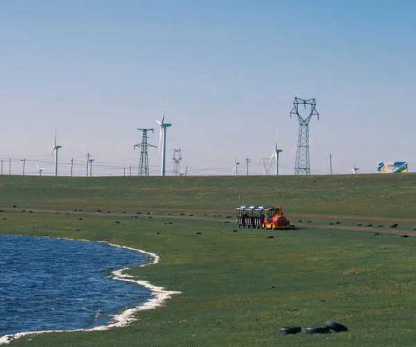 Huitengxile Grassland,Hohhot,Chinatravel Wide coastal plain with a blue lake on the left, wind turbines and transmission towers on the horizon, and an orange maintenance vehicle with passengers near the center.