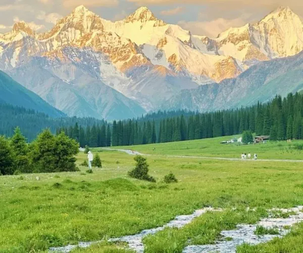 Narati Grassland,Ili,China tour Snow-capped mountains bathed in golden light overlook a green meadow with a winding stream and distant hikers.
