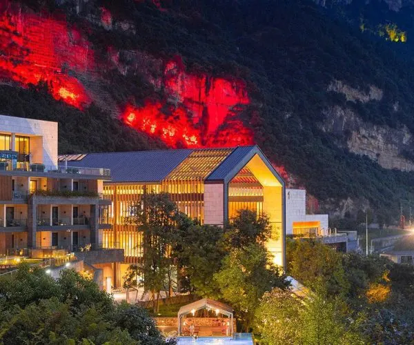 Langjiu Manor,Luzhou,Chinatravel Modern resort buildings at night with warm interior lights, a lit pool area, and a red-lit rocky cliff rising behind.