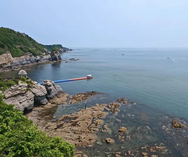 Golden Pebble Beach (Jinshitan) National Geopark,Dalian,China tour Coastal rocky shoreline with a small domed pavilion on a cliff, a colorful floating platform extending into calm blue water, and people exploring the rocks below.