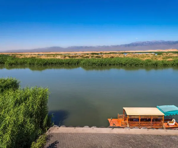 Yinchuan Weather,Yinchuan,Chinatrip Calm river with reed-lined banks, desert dunes and distant mountains under a blue sky; a wooden canopy boat is docked at the shore.