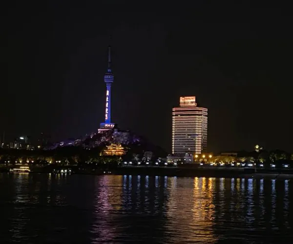 Wuhan Yangtze River Cruise,Wuhan,Chinatravel Night cityscape featuring a tall illuminated tower atop a hill and a striped lit hotel by the river.