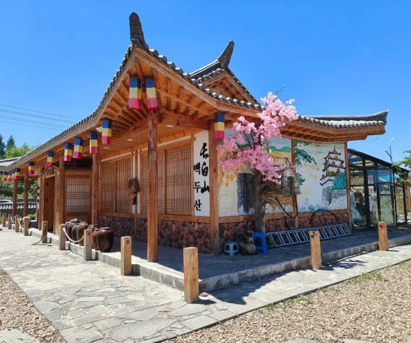 Changbai Korean Folk Village,Baishan,Chinatrip Traditional Korean hanok-style building with wooden beams, colorful lanterns, and a stone-paved path under a bright blue sky, pink blossoms nearby