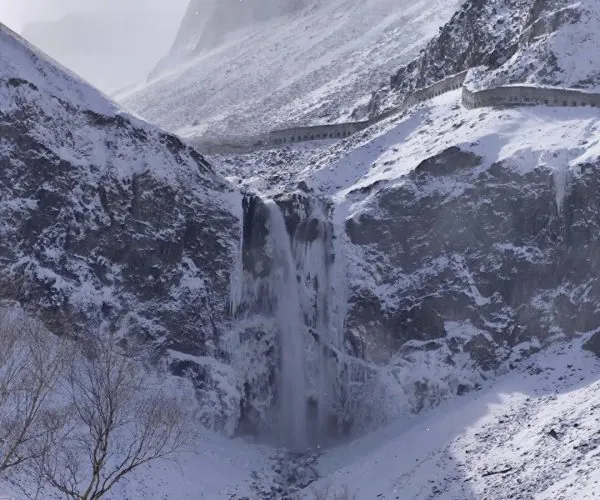 Changbai Waterfall,Baishan,Chinatrip Snowy mountain landscape with a tall waterfall pouring down a rocky cliff, a long curved stone wall snaking along the ridge, and leafless trees in the foreground.