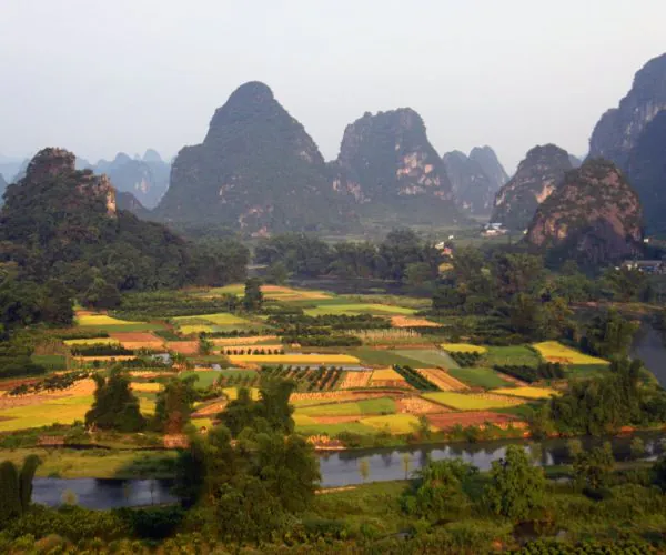 Yangshuo Weather,Yangshuo,Chinatravel Patchwork agricultural fields with a river in the foreground and towering karst mountains in the distance under a hazy sky.