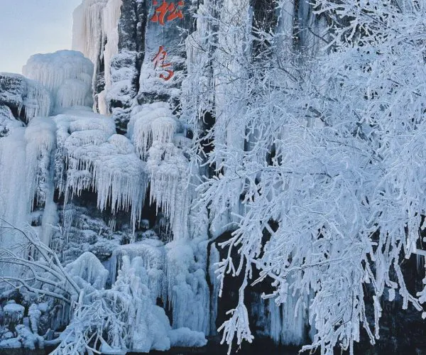 Rime Island,Jilin,Chinatrip Ice-covered rock face with red Chinese characters carved into stone, frozen waterfalls cascading down, snow-laden trees by a calm body of water.