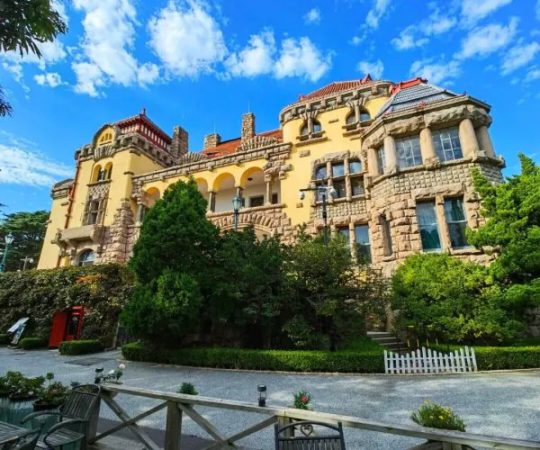 Former German Governor’s Mansion,Qingdao,Chinatravel Castle-like yellow mansion with red-tiled roof and stone accents, framed by trees under a bright blue sky; outdoor seating in the foreground.