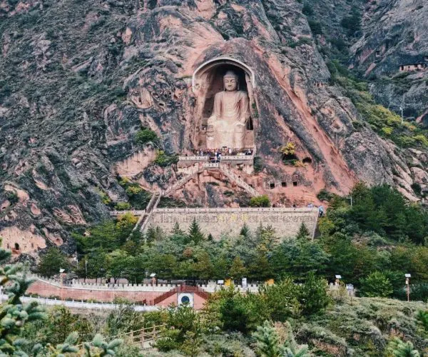 Xumishan Grottoes,Guyuan,China tour Large Buddha statue carved into a rock cliff with surrounding cave alcove, overlooking a temple complex and stairways in a mountainous area