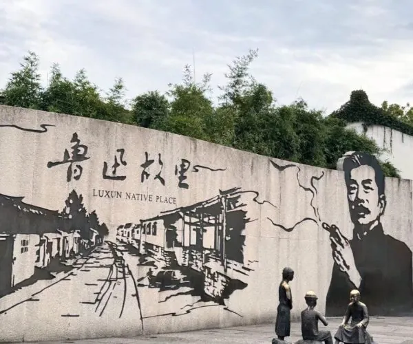 Lu Xun Native Place,Shaoxing,Chinatravel Mural reading 'Luxun Native Place' with a black-and-white village street scene and a large portrait on the right; foreground shows a small sculpture group of four people.