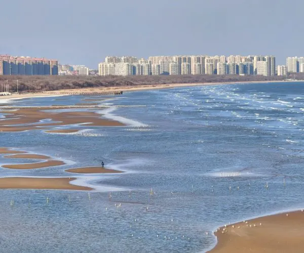 Pigeon Nest Park,Qinhuangdao,Chinatravel Urban skyline along a sandy beach with tidal pools and winding sandbars; a person walks on a sandbar in the foreground.