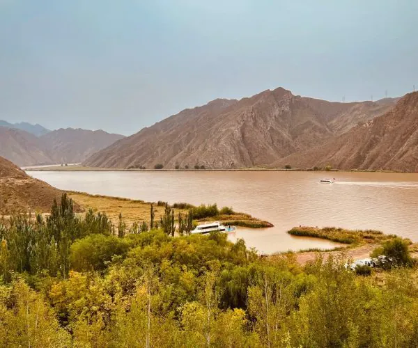 Yellow River Canyon,Zhongwei,China tour Brown river lake with boat cruising near rocky hills and distant mountains, green vegetation in foreground