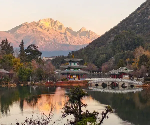 Black Dragon Pool,Lijiang.Chinatrip Tranquil Chinese garden with a tiered pagoda, a stone arch bridge, and a calm lake with mountains in the distance at sunset.