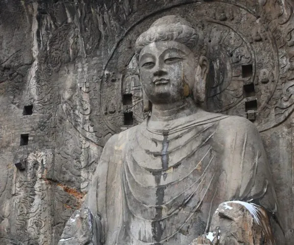 Longmen Grottoes,Luoyang,Chinatravel Rock-cut Buddha statue carved into a cliff with a circular halo behind the head and intricately carved reliefs on the cave wall, with patches of snow on the rock.